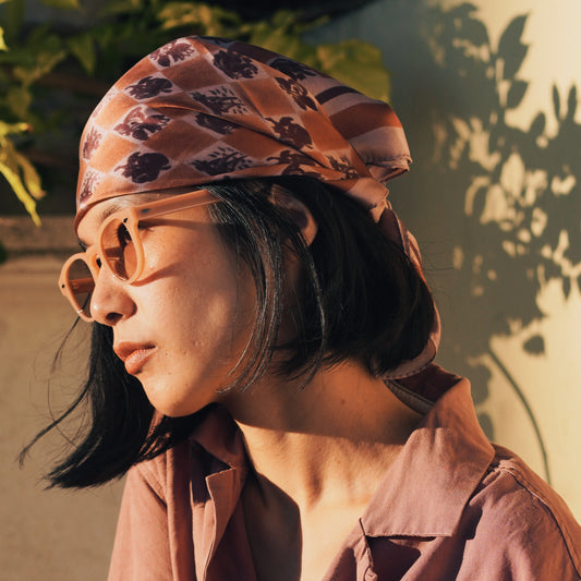 Woman wearing the Santa Luzia pink silk foulard from Tillerman as a headscarf, styled with warm-toned sunglasses and a rose shirt in soft evening light.