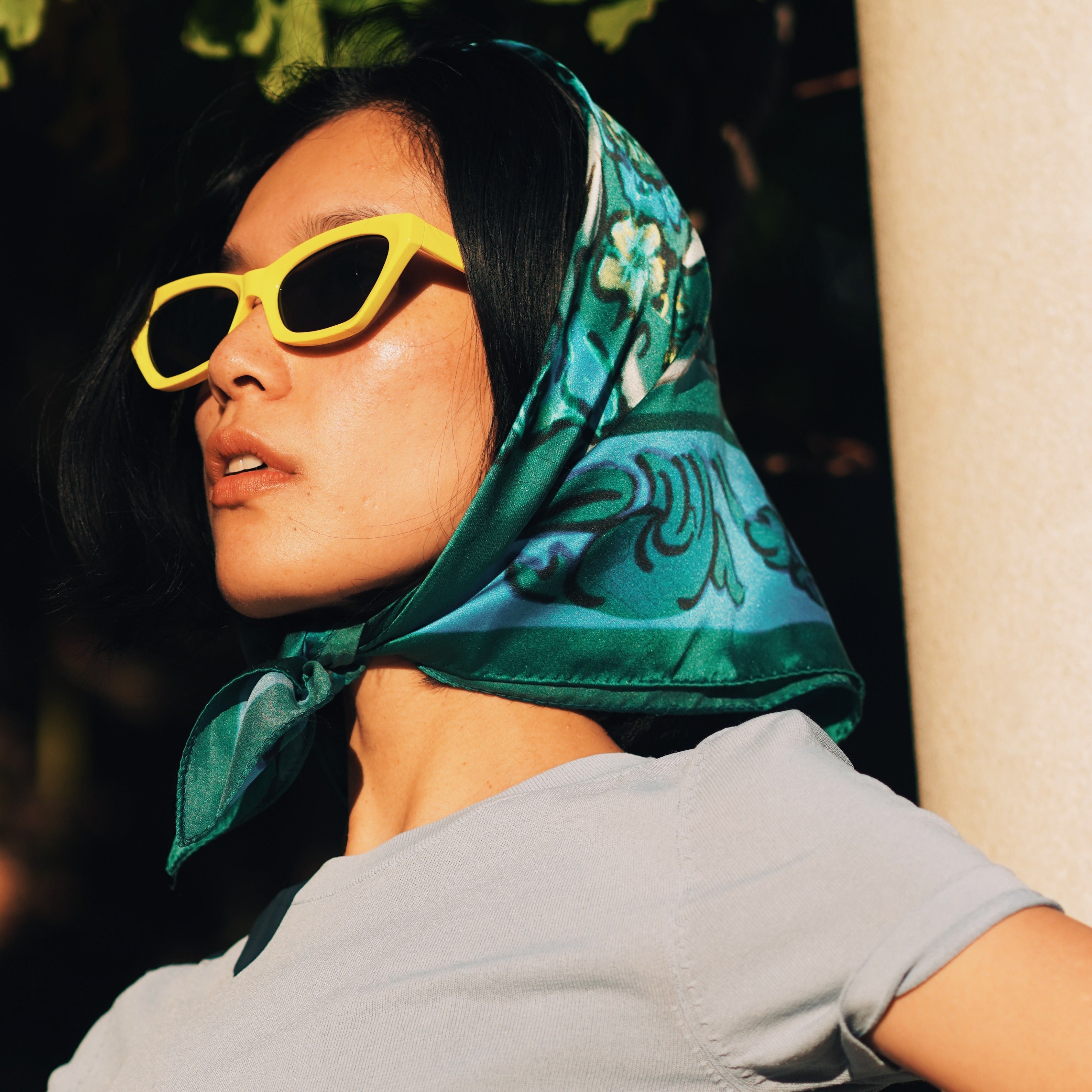 Model wearing the VASO DE FLORES blue silk scarf as a chic headscarf paired with yellow sunglasses – a luxury twill silk accessory by Névoa Zurich inspired by Mediterranean art