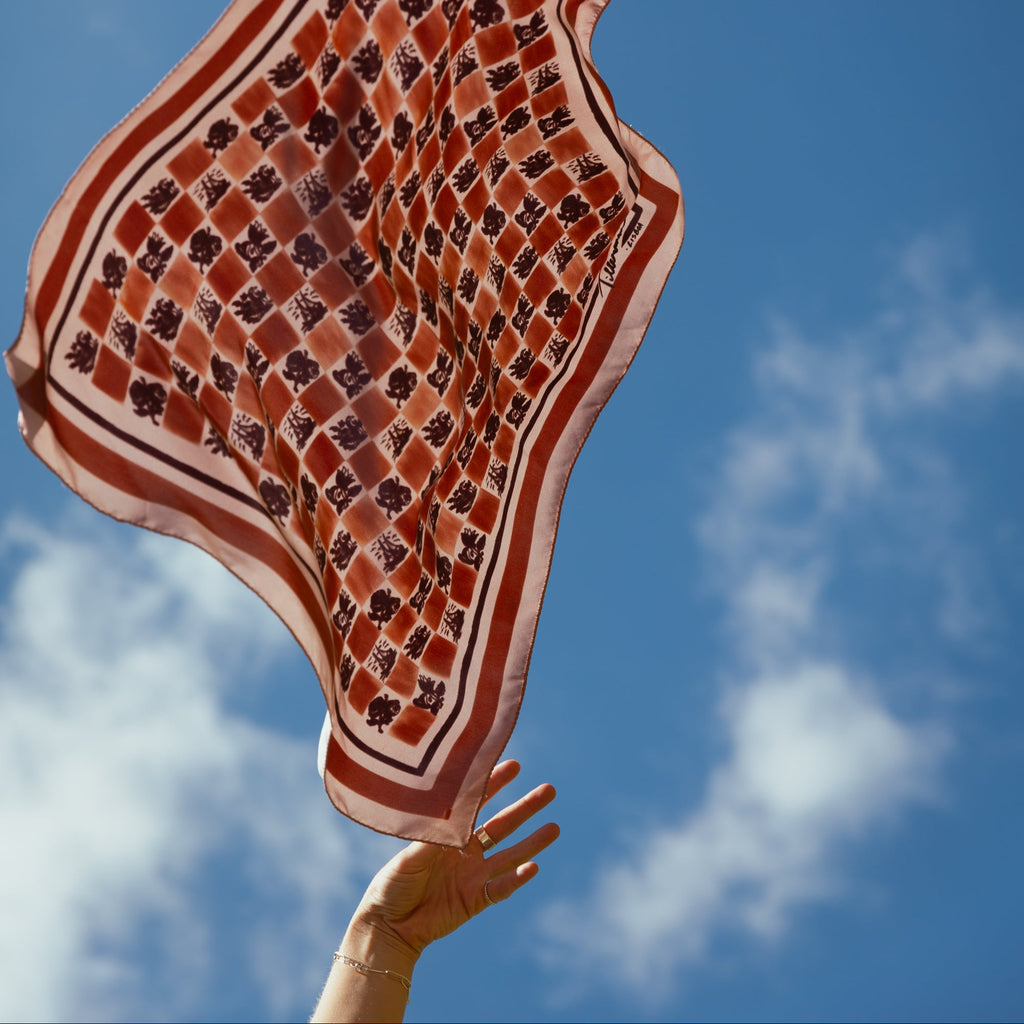 Hand holding the Santa Luzia silk foulard with a floral check pattern against a blue sky, symbolizing lightness and movement — Tillerman x Névoa Zurich.