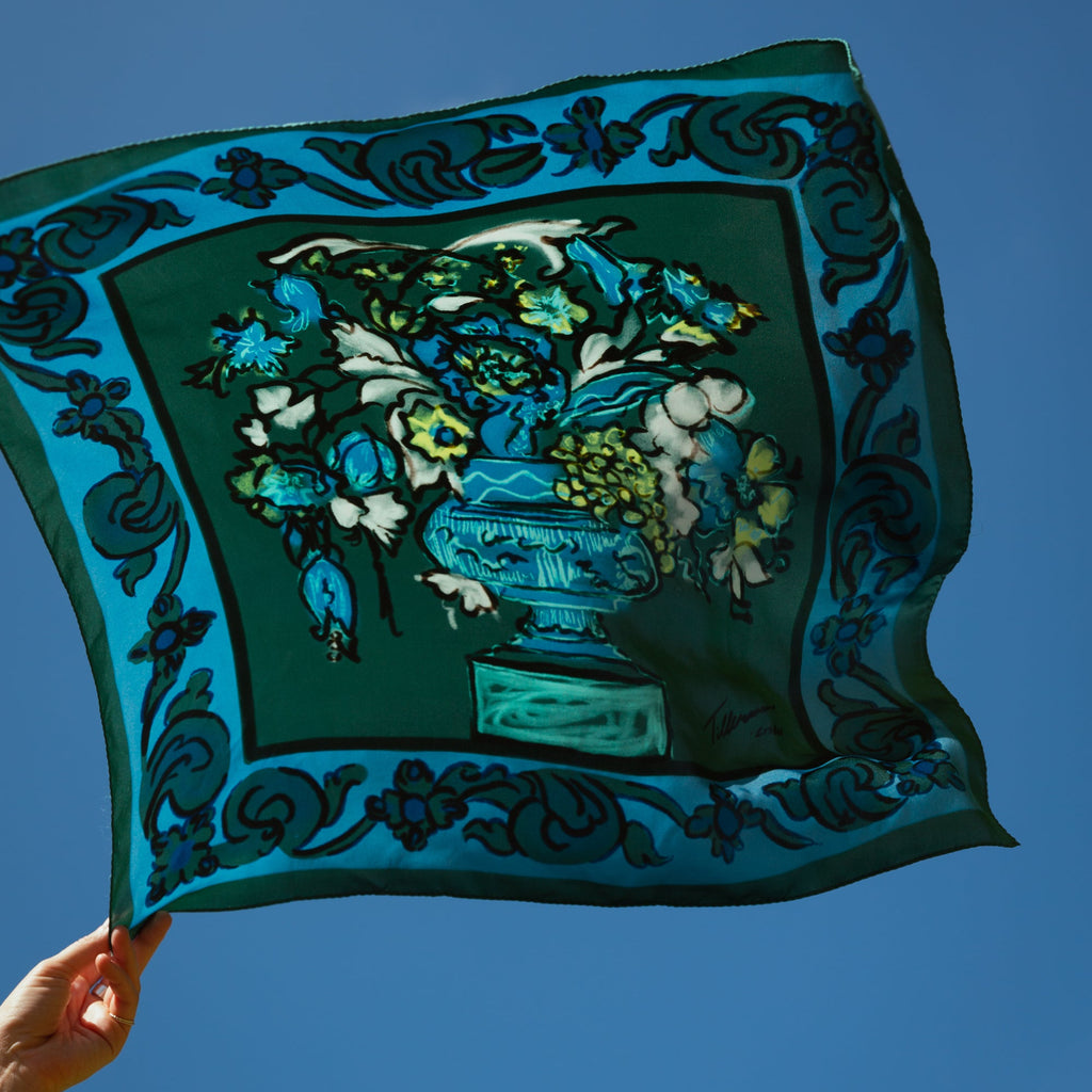 The VASO DE FLORES blue silk scarf by Névoa Zurich, featuring a hand-drawn floral vase motif with ornamental border, held up against a bright blue sky. A Mediterranean-inspired luxury accessory crafted from 100% twill silk