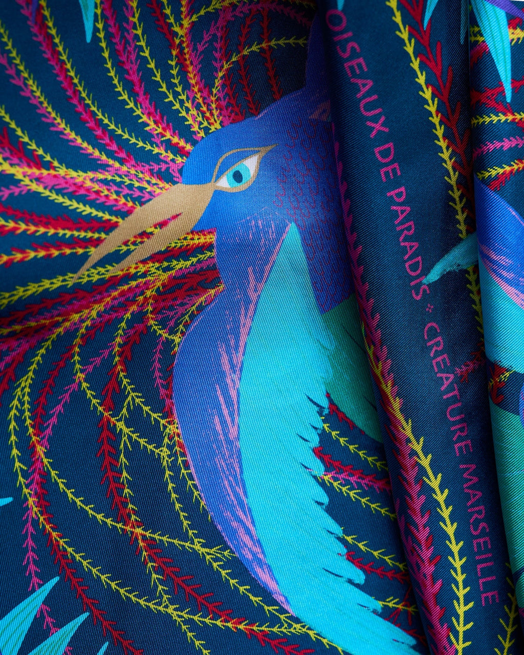 close up view of silk foulard named les oiseaux de parais with a blue, turquoise bird in the center and yellow, red and pink decorative elements