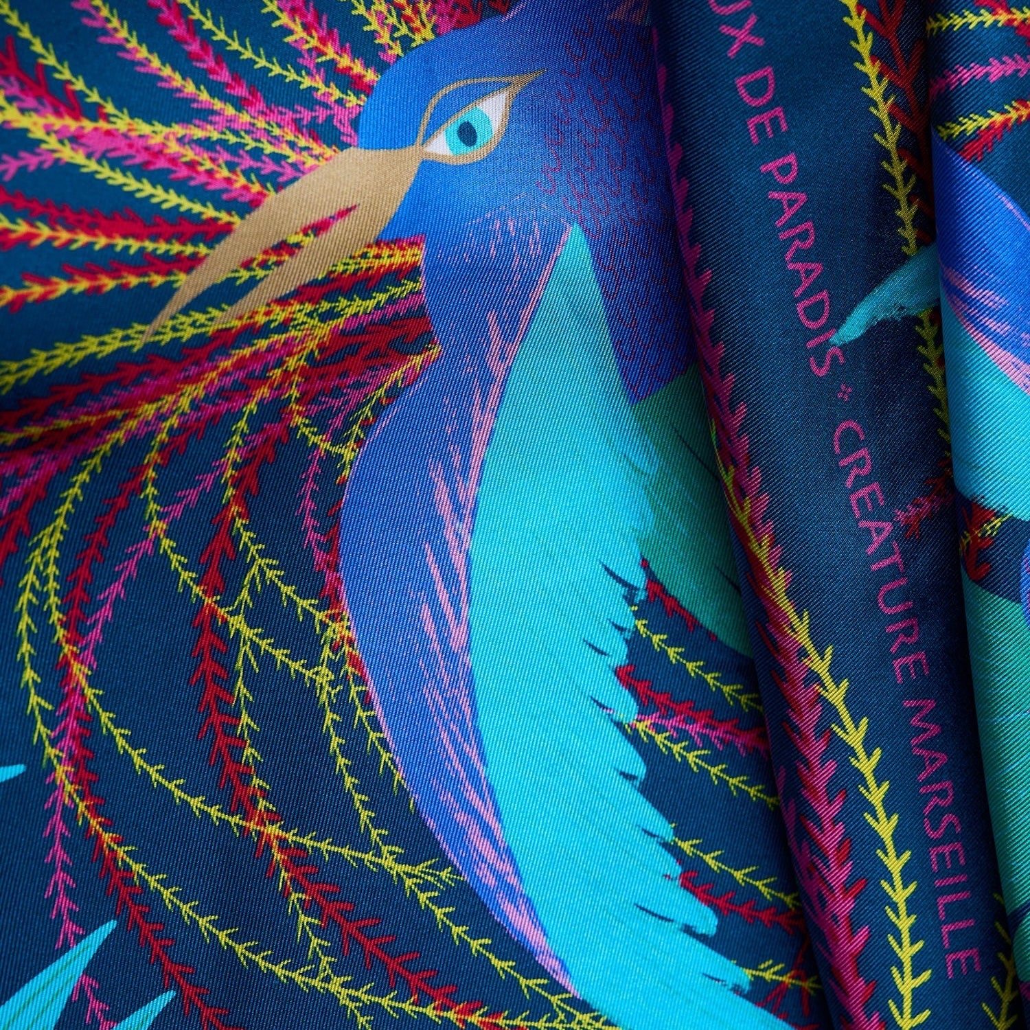 close up view of silk foulard named les oiseaux de parais with a blue, turquoise bird in the center and yellow, red and pink decorative elements